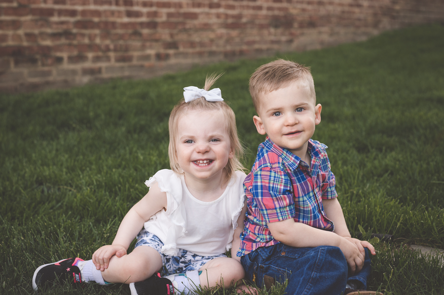 Two toddlers, a girl on the left and a boy on the right sit next to each other on a hill of green grass with a red brick wall in the background. Both children are white and have blue eyes and dark blonde hair. The girl is side siding and is wearing a white bow in her hair, a white shirt with ruffles, blue and white floral shorts, black shoes, and ankle and foot orthotics. She has a big smile on her face and does not yet have all of her baby teeth. The little boy has his back leaned against his sister’s shoulder and is smiling at the camera. He is wearing a blue and pink plaid shirt with jeans and cowboy boots.