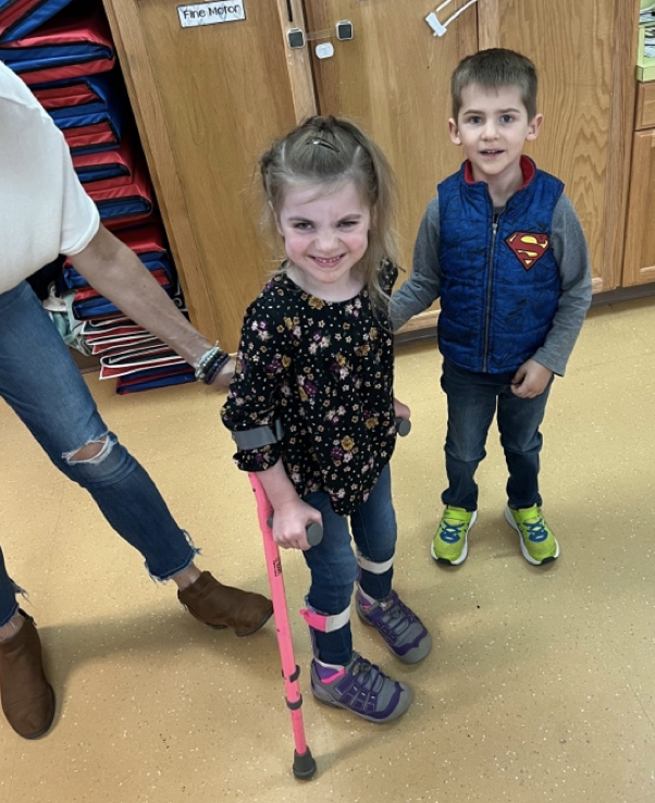 A little girl with light brown hair and blue eyes stands in the middle of the photo. She is wearing a black floral shirt, jeans, and pink and purple ankle and foot orthotics. Both her arms are in bright pink forearm cutches, and she looks very happy about it. A little boy is to the right in the frame. He is wearing jeans, bright yellow trainers, and a blue Superman vest over a gray shirt. He has the same light brown hair and blue eyes as the little girl, and his arm is supporting her. A woman in a white shirt, ripped jeans, and brown boots is also in the background supporting the girl. They are standing in front of a brown cabinet and a stack of blue and red nap mats.
