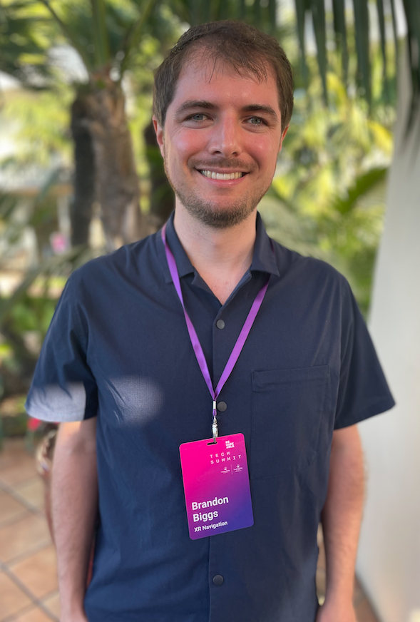 XR Navigation CEO Brandon Biggs outside with palm trees in the background. Smiling white man with green eyes and brown hair. Wearing a navy collared shirt and a purple lanyard that has his nametag on it.
