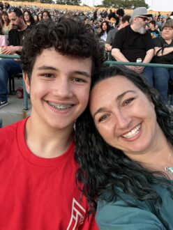 on the left is a smiling boy with brown hair and braces, wearing a red t-shirt with white writing. his smiling mom with dark curly hair leans into him. she's wearing a sea green shirt