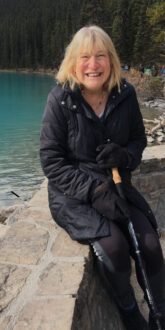 Kim, a white woman with blond hair, wearing an all black outfit and holding a hiking pole, sits on a stone wall with blue water behind her.