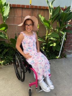 A photo of Miracle, a girl with long brown hair wearing a floppy hat and sundress sitting in her wheelchair