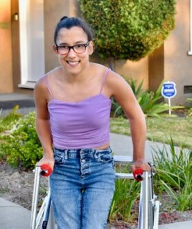 A photo of Miracle, a girl with pulled back brown hair. She is wearing a purple tank top and jeans using her walker.