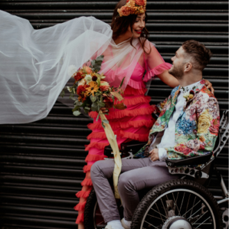 A joyful couple is celebrating outdoors in vibrant, colorful attire. The woman is wearing a bright pink ruffled dress and a floral headpiece, holding a bouquet of sunflowers and red blooms. She smiles down at the man beside her, who is seated in a power wheelchair and wearing a floral-patterned jacket. A flowing white veil adds motion and elegance to the scene.