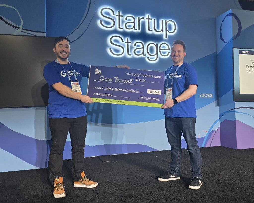 Two people wearing matching blue “Good Trouble” shirts stand on the CES Startup Stage, smiling and holding an oversized check for $20,000 labeled “The Solly Rodan Award.” A large illuminated “Startup Stage” sign is behind them, with CES branding visible on the stage backdrop.
