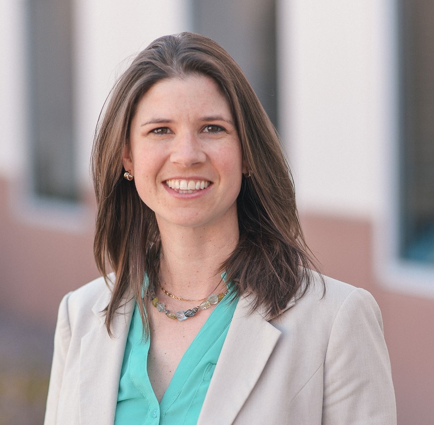 Rhea May Headshot Professional headshot of a smiling woman with shoulder-length brown hair, wearing a light beige blazer over a teal blouse and layered necklaces, standing outdoors with a softly blurred architectural background.